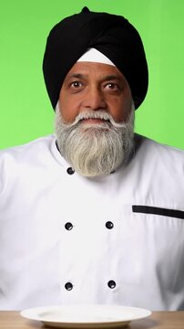 Indian Senior Male Sikh Chef in Traditional Attire Sitting at Table, Pointing at Empty Plate and Showing OK Sign on Green Screen