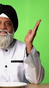 Indian Senior Male Sikh Chef in Traditional Attire Sitting at Table, Pointing at Empty Plate and Showing OK Sign on Green Screen