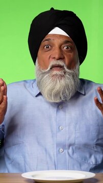 Indian Asian Senior Sikh Man in Casual Clothing Sitting Isolated at Table on Green Screen, Pointing at Empty Plate and Showing OK Sign with Cheerful Expression