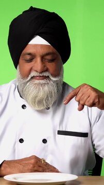 Indian Senior Male Sikh Chef in Traditional Attire Sitting at Table, Pointing at Empty Plate and Showing OK Sign on Green Screen