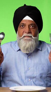 Indian Asian Senior Sikh Man in Casual Clothing Sitting Isolated at Table on Green Screen, Pointing at Empty Plate and Showing OK Sign with Cheerful Expression