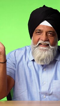 Indian Asian Senior Sikh Man in Casual Clothing Sitting Isolated at Table on Green Screen, Using Mobile App to Order Food with an Empty Plate in Front