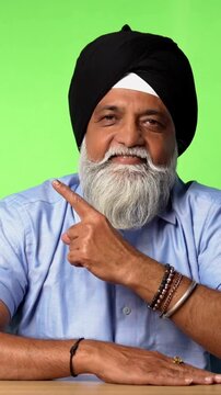 Indian Asian Senior Sikh Man in Casual Clothing Sitting Isolated at Table on Green Screen, Showing Surprise Expressions, Thinking, Thumbs Up, or Pointing at Blank Space
