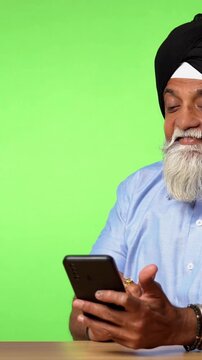 Indian Asian Senior Sikh Man in Casual Clothing Sitting Isolated at Table on Green Screen, Using Smartphone to Surf the Internet and Chat with a Happy Expression