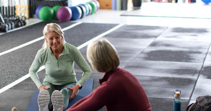 Two women with instructor in green leading warmup showing seated hamstring stretch on mat in gym