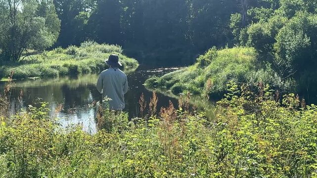 A fisherman with a fishing rod stands in the tall grass on a high riverbank on a sunny, clear summer day. Fisherman on the weekend. Hobbies, recreation and active recreation in summer
