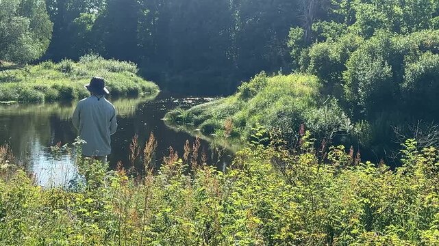 A fisherman with a fishing rod stands in the tall grass on a high riverbank on a sunny, clear summer day. Fisherman on the weekend. Hobbies, recreation and active recreation in summer