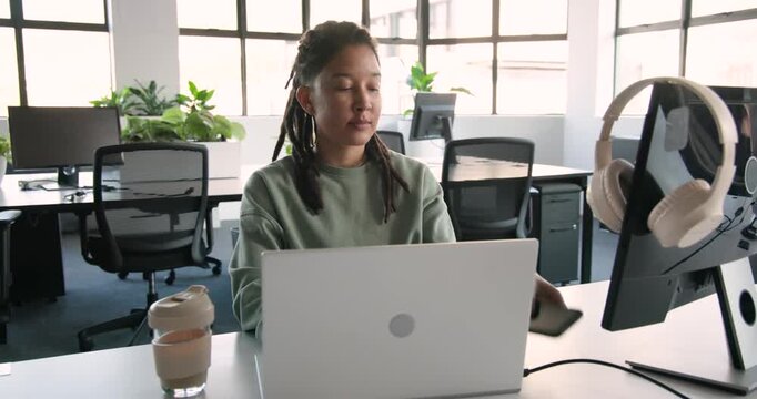 Glancing at monitor, nonbinary worker in green sweater reaching for phone recording message at desk
