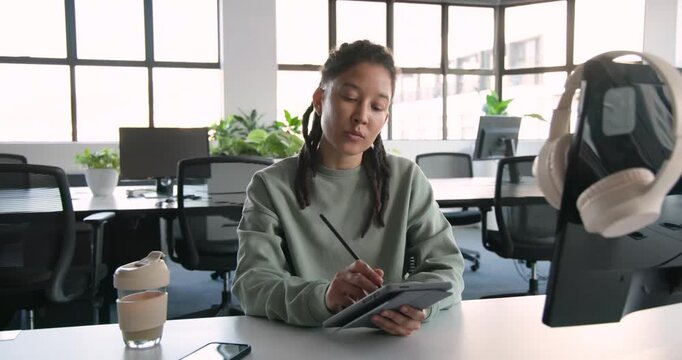 Non-binary African designer holding tablet and sketching with stylus for work at desk, phone cup