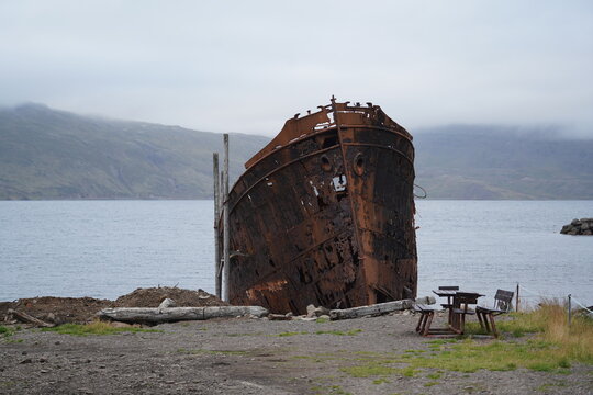 View of the rusted shipwreck of the Sudurland resting on the rocky shore of a fjord under an overcast sky in Djupavik, Arneshreppur, Iceland.