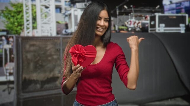 Woman latin holding red heart gift box with bow points thumb over shoulder in studio amid stage equipment smiling at camera; romance surprise.