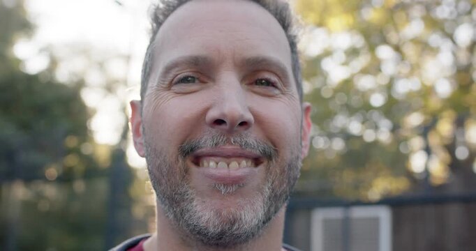 Middle-aged man in maroon tee smiling in backyard by wood fence, lens flare causing smile widening
