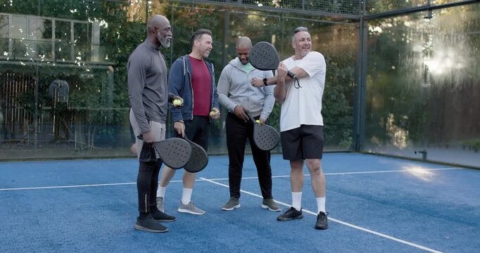 Diverse male friends placing balls on bases, paddles on padel court after white-shirt cue for drill