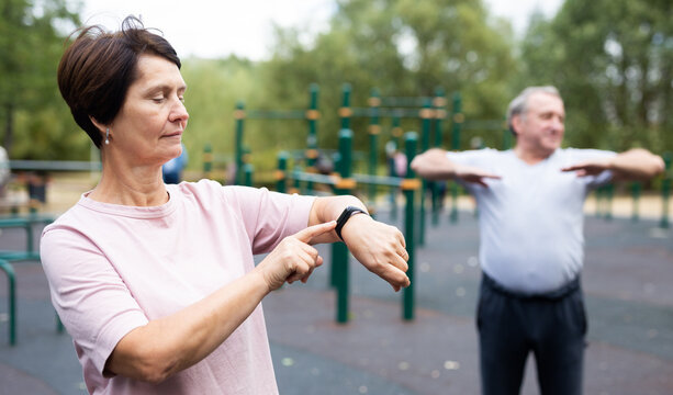 Mature woman in sportswear looks at fitness watch or at pedometer while exercising in city park