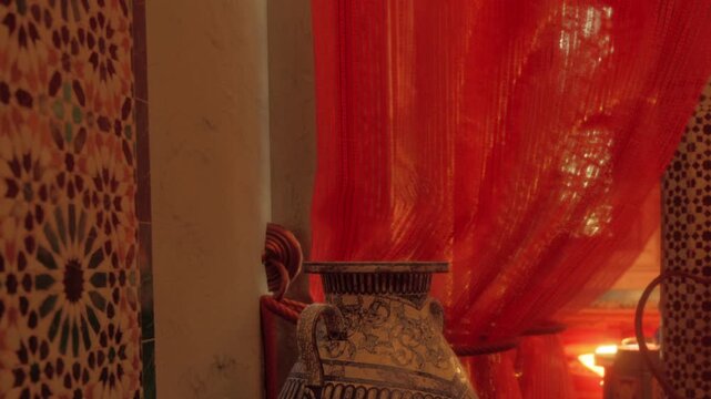 A view of decorative pottery sits in a room with red curtains. The pottery displays intricate designs. Soft light filters through the curtains, enhancing the warm atmosphere.