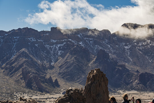View of the volcanic landscape of Mount Teide with snow-capped peaks, rocky formations, and clouds under a blue sky, with tourists in Santa Cruz de Tenerife, Canary Islands, Spain.