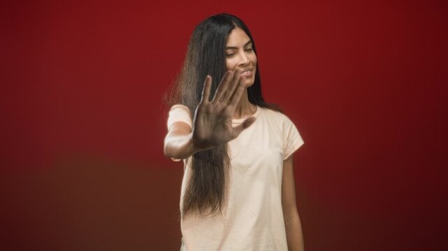 Woman with long hair in a beige t shirt shows palm to camera and mimics steering wheel with both hands in a red studio; caution boundary privacy.