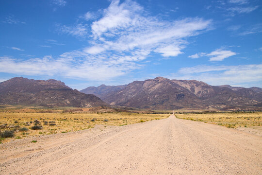 Eine lange Schotterstra&szlig;e verl&auml;uft gerade durch eine trockene Ebene vor dem Brandberg in Namibia.