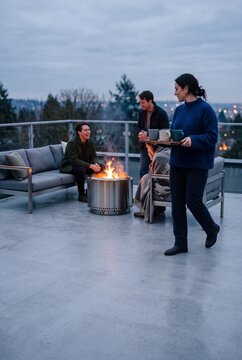 A group of young friends enjoying a cozy autumn evening around a modern fire pit on a rooftop terrace, with a woman bringing hot drinks on a tray