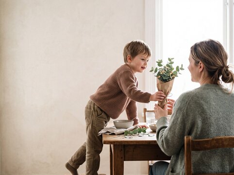 Cute little boy giving a green eucalyptus bouquet to his happy mother while sitting at a rustic wooden table in a bright cozy home