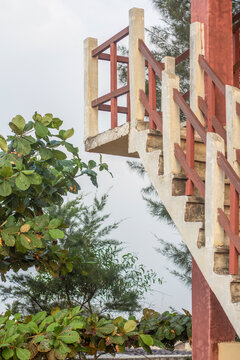 Low-angle concrete stairs on a tower with a red railing, framed by green trees against a gray sky