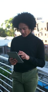 Vertical video: African American man in turtleneck holding tablet, tapping and reading on balcony