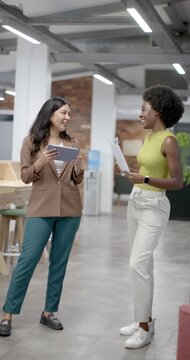 Vertical video: Diverse female coworkers smiling in office, checking tablet and docs for meeting