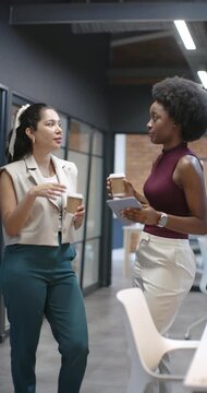 Vertical video: Female coworkers chatting after checking tablet in office corridor with coffee
