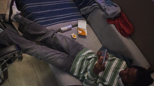 African American teenager lying on couch, playing mobile games on smartphone near junk food, top view. Footage is perfect for social ads addressing screen addiction and teenage mental health