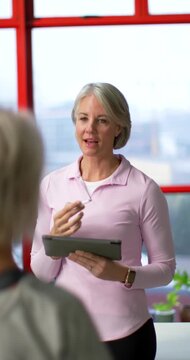 Vertical video: Senior women, coach holding tablet and stylus, tapping to show notes by red window