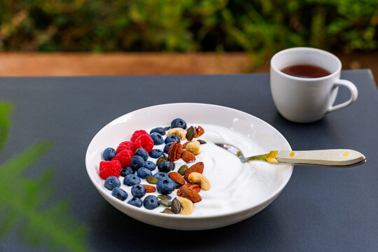 Healthy yogurt bowl topped with fresh blueberries, raspberries, mixed nuts and seeds, served with tea or coffee on table, wellness and breakfast concept
