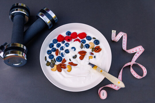 Top view of healthy yogurt bowl with berries and nuts near dumbbells and measuring tape, fitness nutrition and weight loss concept