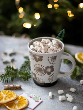 Hot chocolate topped with mini marshmallows and cocoa powder in patterned mug with wooden tree pick, dried orange slices and bokeh lights, close-up