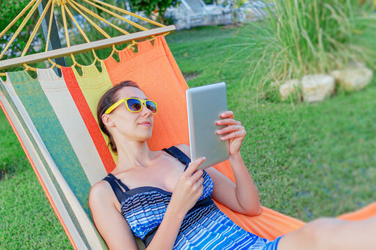 A relaxed woman with yellow sunglasses lies in a colorful hammock while holding a digital tablet in a green garden setting.