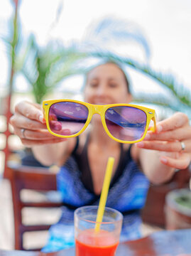 A woman holds up bright yellow sunglasses towards the camera while enjoying a tropical drink outdoors.