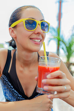 A smiling woman wearing yellow sunglasses holds a glass of red juice outdoors during a sunny day.