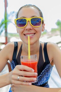A smiling young woman wearing yellow sunglasses drinks a fresh red juice through a yellow straw on a sunny summer day.