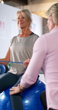 Vertical video: Female senior and trainer touching shoulder and thigh pulling band on ball in rehab