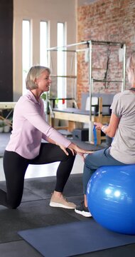 Vertical video: Mature PT placing guiding aligning knee in rehab on ball holding band, copy space