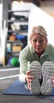 Vertical video: Mature female stretching on blue mat in gym holding shoes, warming up in green top