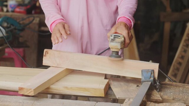 Carpenter's hands using an electric orbital sander to smooth a wooden plank in a woodworking workshop, part of the furniture making and finishing process.	