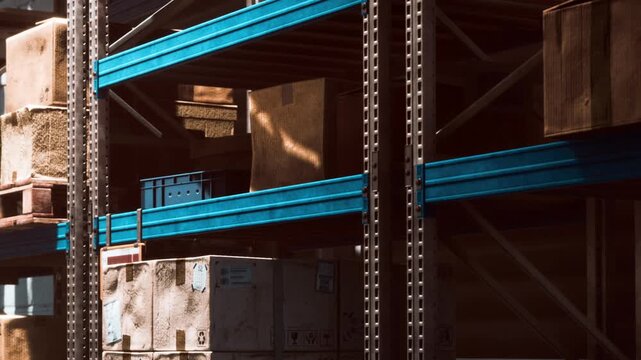 A storage facility shows shelves filled with different boxes and crates. Some items are stacked high while others are placed lower. Light filters through the space and creates shadows.