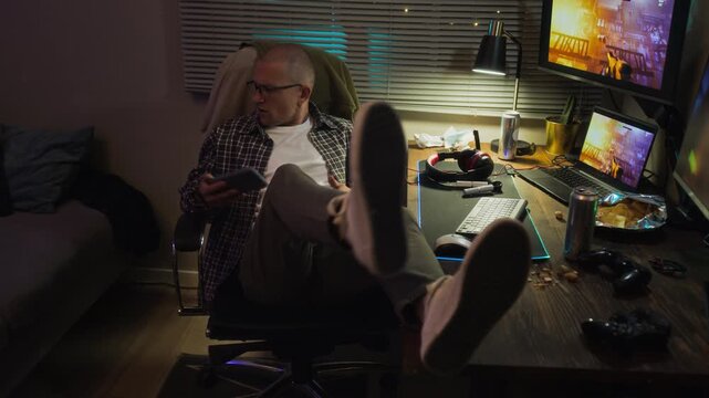 Caucasian young adult man playing mobile game, sitting up to put on headphones and continuing gameplay on computer while drinking soda. Video is perfect for gaming addiction and esports marketing