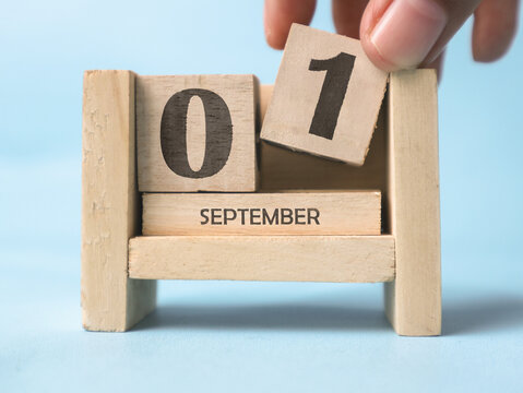 A woman's hand is holding a wooden block with the number 1 written on it on a wooden block calendar for September 1st on a light blue background.
