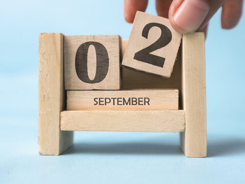 A woman's hand is holding a wooden block with the number 2 written on it on a wooden block calendar for September 2nd on a light blue background.