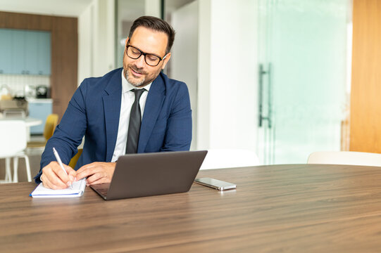 Professional male entrepreneur concentrating on business, writing notes in a notebook while utilizing a laptop, managing finances, bookkeeping, and legal documentation