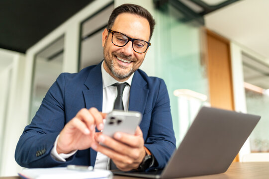 Smiling professional businessman interacting with a mobile phone, managing tasks and digital communication from his office desk with a laptop open