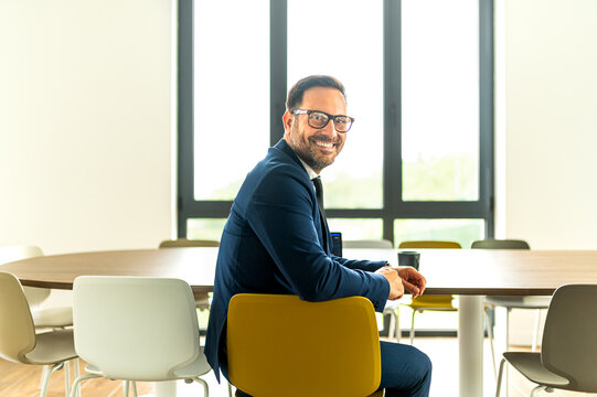 Smiling entrepreneur wearing glasses, sitting at a conference table with a coffee cup and laptop, ready for a productive discussion or collaboration in a modern workplace setting