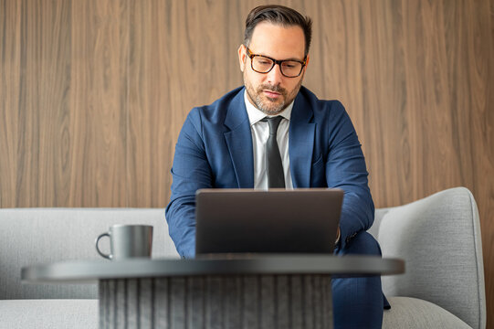 Entrepreneurial man in business attire sitting on a gray couch, analyzing data on a portable computer with a coffee cup nearby, actively engaged in online tasks