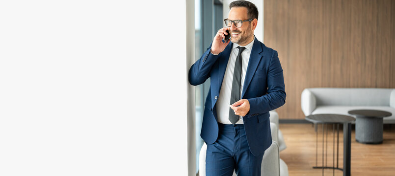 Businessman communicating on a mobile phone, smiling while standing in a modern office, discussing important business details, managing finances, and consulting clients